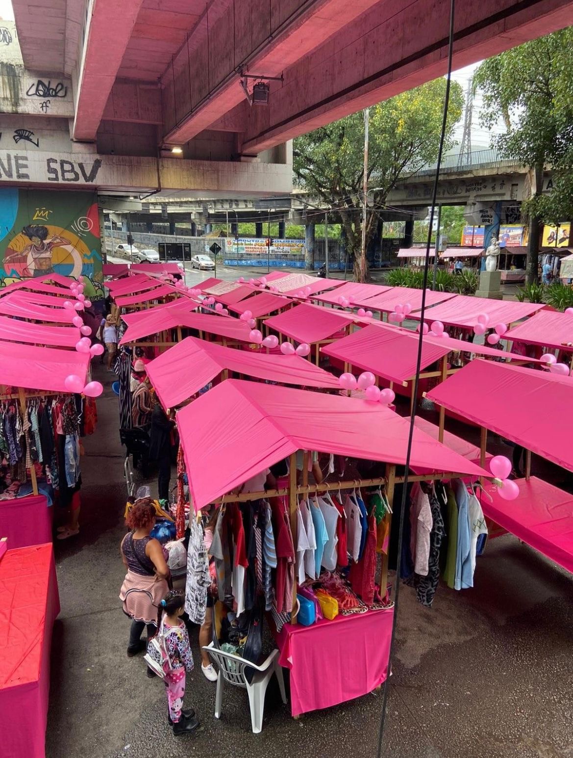 Barracas rosas da Feira de Brechó Cachorrão embaixo do viaduto, com araras de roupas e balões